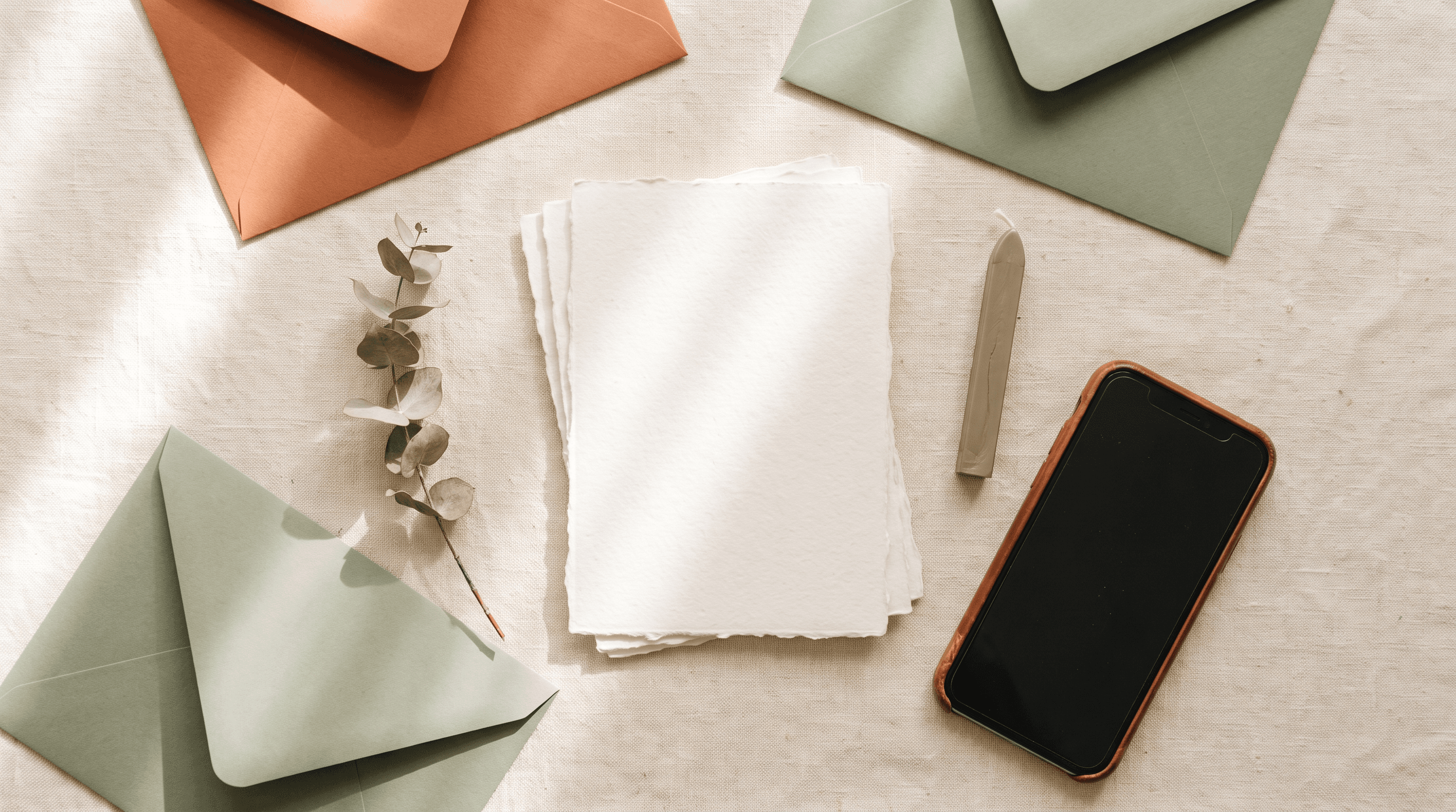 Blanco trouwkaarten met terracotta en saliegroene enveloppen naast een smartphone op een linnen tafelkleed